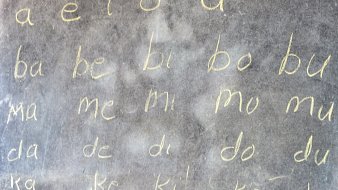 L'image montre un tableau noir sur lequel sont écrits des caractères en lettres cursives, à la craie. Les lignes commencent par les voyelles "a", "e", "i", "o", "u", suivies de combinaisons de consonnes et voyelles, telles que "ba", "be", "bi", "bo", "bu", puis "ma", "me", "mi", "mo", "mu", et ainsi de suite. L'écriture est claire et simple, et le tableau a une texture légèrement rugueuse. L'ensemble de l'image évoque un environnement d'apprentissage.