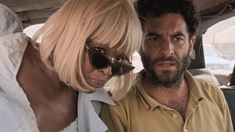 Dans cette image, on voit deux personnes assises à l'arrière d'un véhicule. À gauche, une femme d'une trentaine d'années a les cheveux blonds et courts, coiffés en une coupe élégante. Elle porte de grandes lunettes de soleil noires et une tenue légère. Son expression est attentive, presque inquisitrice. À droite, un homme d'une trentaine d'années également, a une barbe foncée et des cheveux bouclés. Il est vêtu d'une chemise de couleur terre. Son regard est préoccupé et intense, comme s'il était en train de réfléchir à quelque chose d'important. L'atmosphère dans l'image semble suggérer une conversation sérieuse ou un moment de tension partagée. Les sièges du véhicule sont en cuir ou en tissu sombre, et on peut apercevoir des objets non identifiables, peut-être des sacs ou des vêtements, à leurs pieds.