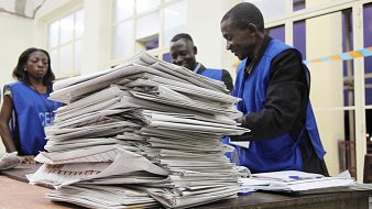 L'image montre une scène à l'intérieur d'un bureau de vote. Au premier plan, on aperçoit une grande pile de documents, probablement des bulletins de vote ou des formulaires électoraux, soigneusement empilés sur une table en bois. À l'arrière-plan, trois personnes en gilets bleus se trouvent près de la table, semblant discuter ou examiner les papiers. L'atmosphère est concentrée, suggérant une activité importante liée à un scrutin. La lumière éclaire la pièce, créant une ambiance sérieuse et professionnelle.