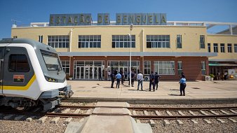 Cette image montre la gare de Benguela, un bâtiment aux couleurs pâles et au style architectural distinctif. À l'avant, on voit un train moderne garé sur une voie ferrée. L'architecture de la gare est caractérisée par des fenêtres spacieuses qui laissent entrer beaucoup de lumière naturelle. Devant la gare, un groupe de personnes, probablement des voyageurs ou du personnel, se tient debout, discutant ou attendant. Le ciel est clair et ensoleillé, créant une ambiance vive et accueillante. Les rails de la gare sont visibles au premier plan, ajoutant au contexte ferroviaire de la scène.