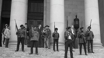 L'image montre un groupe de personnes sur les marches d'un bâtiment imposant, probablement une institution gouvernementale. Au premier plan, plusieurs jeunes hommes se tiennent debout, portant des vestes sombres et des armes à feu, avec des expressions déterminées sur leurs visages. Ils sont entourés d'autres individus, certains vêtus de manière formelle, comme des costumes et des chapeaux. Tous semblent engagés dans une sorte de manifestation ou de rassemblement, émettant un sentiment de tension et de détermination. L'architecture du bâtiment est majestueuse, avec de grandes colonnes en arrière-plan. L'atmosphère générale de l'image évoque une période de lutte pour les droits civiques.