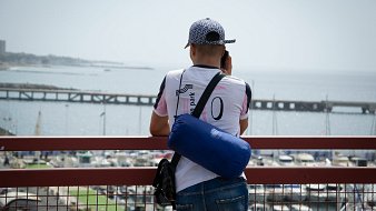 Dans cette image, un homme se tient de dos, regardant au loin vers la mer. Il est vêtu d'un t-shirt blanc avec des accents colorés et porte une casquette à motifs. Sur son épaule, il a un petit sac bleu. L'arrière-plan montre un port avec des bateaux amarrés et une jetée qui s'étend dans l'eau, créant une atmosphère tranquille. Le ciel est clair, suggérant une journée ensoleillée. L'homme semble concentré sur ce qu'il observe, profitant de la vue sur l'horizon maritime.