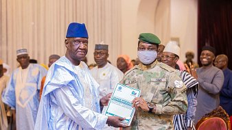 L'image montre un événement officiel ou une cérémonie de remise de prix. Au premier plan, un homme portant une tenue traditionnelle, composée d'une longue robe et d'un chapeau bleu, reçoit un document d'un officier militaire. Ce dernier est vêtu d'un uniforme camouflé et porte un masque gris. En arrière-plan, plusieurs personnes en tenue variée assistent à l'événement, certaines applaudissant, tandis que d'autres observent. L'environnement semble formel, avec des décorations adaptées à l'occasion.