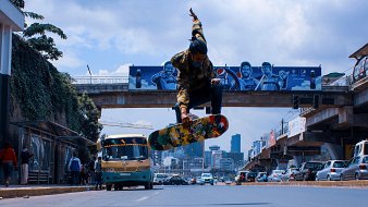 L'image montre un skateur en plein saut au-dessus de la route. Il est habillé d'un sweat à capuche avec des motifs et porte un skateboard coloré. En arrière-plan, il y a des bâtiments urbains, des graffitis sur un mur, et un bus qui passe, typique des grandes villes. La scène capture une ambiance dynamique et énergique, avec un ciel partiellement nuageux qui ajoute à l'atmosphère urbaine. Les paysages urbains et la présence d'autres personnes montrent l'animation de la ville.