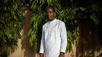 L'image montre un homme debout devant un arrière-plan de feuillage vert dense. Il porte une tunique blanche à col montant et un pantalon sombre. Son sourire dégage une attitude amicale et confiante. Les arbres autour de lui ajoutent une ambiance naturelle, avec des branches feuillues qui encadrent sa silhouette. Le fond est constitué de murs dans des tons nude, apportant une touche chaleureuse à la scène. La lumière naturelle semble douce, créant une atmosphère sereine.