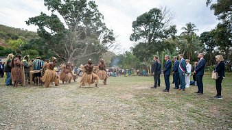 L'image montre un groupe de personnes en tenue traditionnelle, qui semblent participer à une cérémonie ou une célébration. Au centre, des hommes vêtus de costumes en fibres naturelles dansent avec énergie. Ils sont entourés par un public, composé de personnes en tenue formelle, qui observe attentivement la scène. L'environnement est verdoyant, avec des arbres et une légère brume qui donne une atmosphère presque mystique. À l'arrière-plan, un groupe de spectateurs semble apprécier l'événement, ajoutant une dimension communautaire à cette célébration culturelle.