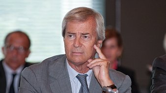 L'image montre un homme d'âge moyen, assis à une table de conférence. Il a les cheveux gris et porte un costume gris. Son expression est pensive, et il touche son visage avec un doigt en réfléchissant. En arrière-plan, on peut distinguer d'autres personnes assises, mais leurs visages sont flous. L'ambiance semble professionnelle, suggérant une réunion ou une discussion importante. La lumière est douce, créant une atmosphère de concentration.