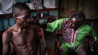 L'image montre deux hommes assis dans des chaises en bois dans une pièce intérieure. L'un des hommes, assis à gauche, a un torse nu et semble pensif. Son expression montre une certaine vulnérabilité. L'autre homme, à droite, est vêtu d'une robe de couleur verte et violette, et il semble s'adresser au premier avec attention, posant une main réconfortante sur son épaule. La lumière dans la pièce est douce, créant une atmosphère intimiste. En arrière-plan, on peut voir des objets flous disposés sur des tables, ce qui suggère un environnement familier et intime.