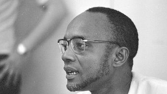 L'image représente un homme portant des lunettes à monture fine. Il a un visage allongé avec une petite barbe bien taillée. Ses cheveux sont courts et légèrement bouclés. L'homme semble concentré et engagé dans une conversation. En arrière-plan, on peut distinguer une silhouette floue d'une autre personne, suggérant qu'il se trouve dans un environnement social ou professionnel. L'éclairage est doux, ce qui met en valeur les traits de son visage.