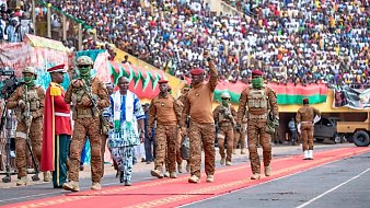 L'image dépeint une scène d'événement public dans un stade. Un grand nombre de personnes est rassemblé dans les gradins, créant une ambiance festive et patriotique. Au premier plan, des militaires en uniforme brun avancent sur un tapis rouge, symbolisant une cérémonie officielle. L'un des militaires, portant une casquette rouge, lève le bras en signe de salutation. À ses côtés, un homme en vêtements traditionnels aux motifs colorés marche avec assurance. La scène est remplie de drapeaux et de banderoles qui ajoutent des touches de couleur à l'événement. L'atmosphère est vibrante, marquée par l'engagement et l'unité.