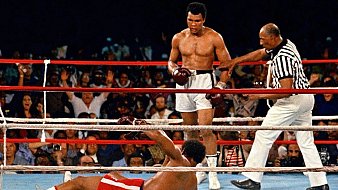 L'image montre un moment mémorable d'un match de boxe. Au centre, un boxeur, torse nu et portant des shorts blancs, se tient debout avec un regard déterminé. C'est Muhammad Ali, connu pour sa rapidité et sa technique. À ses pieds, un autre boxeur est allongé sur le sol, faisant face au ring, après avoir été mis au tapis. Le public en arrière-plan est en émoi, certains levant les bras en signe d'enthousiasme ou d'étonnement. Un arbitre, vêtu d'un maillot à rayures, s'approche du boxeur au sol, prêt à commencer le compte. L'atmosphère est chargée d'intensité et d'adrénaline, typique des combats de boxe.
