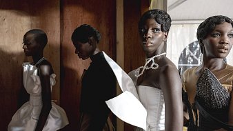 L'image présente un groupe de mannequins lors d'un événement de mode. Les mannequins, d'origines diverses, portent des tenues élégantes et extravagantes, mettant en valeur des tissus texturés et des coupes audacieuses. Au premier plan, l'un des mannequins se distingue par une robe blanche ornée d'un grand nœud, tandis que les autres vêtus de créations plus sombres, affichent des coiffures soignées. La lumière joue sur les tissus, créant un contraste entre les ombres et les zones éclairées, ce qui ajoute une profondeur visuelle à la scène. L'ambiance est à la fois chic et artistique, capturant l'énergie dynamique du monde de la mode.