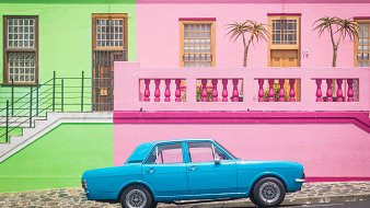 L'image présente une scène vibrante et colorée d'une rue typique. À gauche, un bâtiment se dresse en vert clair, avec des fenêtres en bois et un accès par escalier. À droite, un mur rose vif forme la façade d'une maison, ornée de deux palmiers qui ajoutent une touche tropicale. Entre les deux bâtiments, une voiture de couleur bleu clair est garée, se détachant joliment sur le fond coloré. Le sol est pavé, et l'ambiance générale de la scène est joyeuse et accueillante, évoquant une atmosphère estivale et ensoleillée.