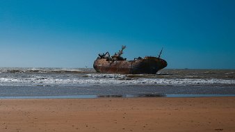 L'image montre un vieux navire échoué sur une plage. Le bateau, rouillé et usé par le temps, repose partiellement immergé dans les vagues calmes de l'océan. En arrière-plan, le ciel est d'un bleu éclatant, sans nuages, tandis que les vagues viennent s'écraser doucement sur le sable doré de la plage. L'atmosphère est à la fois tranquille et mélancolique, évoquant une histoire de voyages passés et d'abandon. Sur la droite, on peut apercevoir quelques oiseaux volant au-dessus de l'eau.