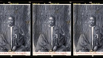 L'image présente trois portraits en noir et blanc d'un homme en costume traditionnel. Il est assis sur un fond de paille tressée qui évoque un environnement naturel. L'homme porte une tenue ample, probablement en textile sombre, et est décoré de plusieurs médailles accrochées à sa poitrine, ce qui suggère un statut élevé ou une reconnaissance. Son visage présente une expression solennelle et sérieuse. Les portraits sont disposés côte à côte, créant une composition symétrique. Cette image témoigne d'un moment historique, probablement lié à une exposition ou un événement marquant.