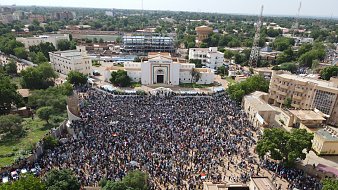 L'image montre une grande foule rassemblée dans une place publique, probablement lors d'une manifestation ou d'un événement collectif. Les personnes sont regroupées en masse, et on peut imaginer une ambiance vibrante, avec des slogans ou des chants. En arrière-plan, des bâtiments se dressent, notamment un bâtiment avec une architecture marquée. Les arbres entourent la zone, ajoutant une touche de verdure. L'atmosphère semble dynamique, illustrant l'engagement de la communauté dans une cause.