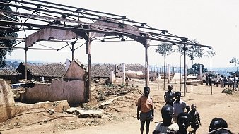 L'image montre un paysage de dévastation avec des structures en ruine. À l'avant-plan, plusieurs personnes, dont des enfants et des adultes, se tiennent debout sur un sol poussiéreux. En arrière-plan, on voit des bâtiments partiellement effondrés, avec des toits manquants et des murs endommagés. Des arbres isolés se dressent ici et là, ajoutant une touche de verdure à ce cadre désolé. Le ciel est dégagé et ensoleillé, contrastant avec l'état des lieux. L'atmosphère suggère une scène de désolation, peut-être après un événement tragique.