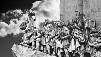 L'image représente un relief sculpté monumentale. On y voit une procession de figures humaines, probablement des explorateurs ou des conquérants, qui se tiennent debout sur une plateforme. Les personnages sont vêtus de costumes historiques et affichent des expressions déterminées. Certains d'entre eux portent des armes, des drapeaux et d'autres objets symboliques, témoignant de leurs exploits. En arrière-plan, on aperçoit des nuages sombres qui ajoutent une atmosphère dramatique à la scène. Le tout est sculpté dans un matériau clair, probablement de la pierre, permettant de bien contraster avec le ciel nuageux. Cette œuvre dégage une impression de puissance et de grandeur, évoquant une célébration de l'histoire et de l'aventure.