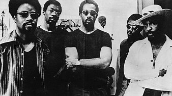 L'image montre un groupe de cinq hommes, tous portant des lunettes de soleil. Ils dégagent une certaine confiance et sont disposés côte à côte. Les hommes sont vêtus de vêtements variés, allant de t-shirts à une chemise blanche large. En arrière-plan, on peut voir des œuvres d'art abstraites et des dessins, ajoutant une dimension artistique au décor. L'ensemble de la scène dégage une atmosphère d'unité et de force, évoquant un moment important ou une rencontre significative.