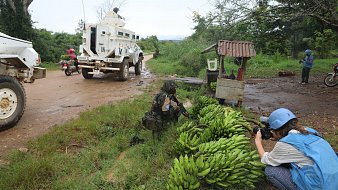 L'image dépeint une scène rurale dans un environnement naturel. Sur le côté gauche, on aperçoit un groupe de bananes bien rangées au sol, d'un vert vibrant. À proximité, un soldat en uniforme utilise une caméra pour capturer des images, se penchant légèrement vers les bananes. Sur la droite, un véhicule blindé est garé sur une route poussiéreuse, entouré d'une végétation luxuriante. Au loin, d'autres personnes, comme un homme sur une moto et une femme à pied, ajoutent une touche de vie à cette scène. Le ciel semble nuageux, suggérant un temps humide, et l'atmosphère générale évoque un mélange d'activités militaires et de vie quotidienne.