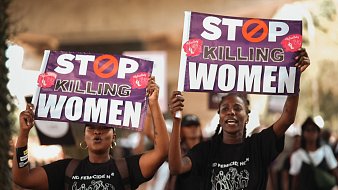L'image montre deux femmes participante à une manifestation. Elles tiennent chacune une grande pancarte. Les pancartes portent le message "STOP KILLING WOMEN" en lettres capitales de couleur blanche, avec un symbole barré en rouge. Les femmes ont des expressions déterminées et sont vêtues de t-shirts noirs. L'arrière-plan suggère une ambiance de mobilisation avec d'autres personnes qui marchent et participent à l'événement. L'atmosphère est chargée d'émotion et de solidarité.