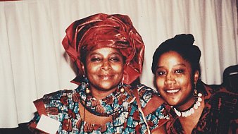 L'image montre deux femmes souriantes posant côte à côte. La femme à gauche porte un vêtement traditionnel aux motifs colorés, accompagné d'un foulard assorti. Sa expression est douce et chaleureuse. À sa droite, la seconde femme affiche également un grand sourire ; elle porte un haut à motifs et des accessoires qui ajoutent une touche élégante à son apparence. L'environnement semble être festif, avec une nappe blanche en arrière-plan, laissant planer une atmosphère accueillante et joyeuse.