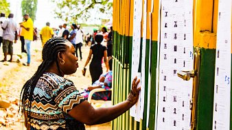Dans cette image, on voit une scène animée en extérieur. Une femme, avec des tresses et portant un haut aux motifs colorés, se tient près d'un mur recouvert de plusieurs feuilles de papier. Elle passe sa main sur l'une de ces feuilles, qui semble contenir des informations, probablement des listes ou des résultats. En arrière-plan, de nombreuses autres personnes se déplacent, créant une atmosphère de rassemblement. Les couleurs vives du mur et la lumière du jour ajoutent une ambiance chaleureuse à la scène. Au sol, on peut apercevoir des feuilles mortes, ce qui suggère une saison automnale.