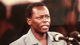 L'image montre un homme en train de prononcer un discours devant un microphone. Il porte une chemise de couleur bleue et semble sérieux et engagé dans ses propos. Son visage exprime une certaine détermination, et il se tient droit. Il y a plusieurs microphones devant lui, et un public peut être présent en arrière-plan, bien que non visible sur cette image. L'atmosphère donne l'impression d'un moment important lors d'un discours ou d'une déclaration publique.