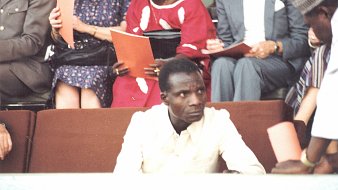 L'image montre une scène d'un événement où un homme en chemise claire est assis au premier plan, semblant sérieux et attentif. À l'arrière-plan, plusieurs personnes discutent, certaines tenant des documents ou des livrets. On aperçoit des vêtements colorés, peut-être d'origine africaine, ce qui ajoute une dimension culturelle à la scène. L'atmosphère générale évoque un rassemblement formel ou une cérémonie.