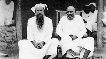 L'image montre deux hommes assis côte à côte sur un tapis au sol. L'homme de gauche, portant une longue barbe blanche et un turban, est habillé d'une tunique blanche. À sa droite se trouve un autre homme, également en tunique blanche, qui semble plus âgé, avec une barbe grise. Ils affichent tous deux une expression calme et sereine. En arrière-plan, on peut distinguer des silhouettes d'autres personnes floues et un environnement rural. Ces détails donnent une impression de tranquillité et de tradition.