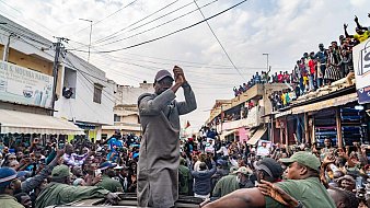 L'image montre une scène dynamique et animée dans une rue bondée. Au centre, un homme, vêtu d'une tenue grise, se tient debout sur un véhicule, levant les bras avec enthousiasme. Les gens autour de lui, une foule importante, applaudissent et expriment leur soutien. On peut entendre des cris de joie et des chants de la part des personnes qui entourent le véhicule. À l'arrière-plan, des bâtiments et des personnes sur les balcons ajoutent à l'atmosphère de ferveur et de célébration. Le ciel est partiellement nuageux, créant un éclairage intéressant sur cette scène vibrante.