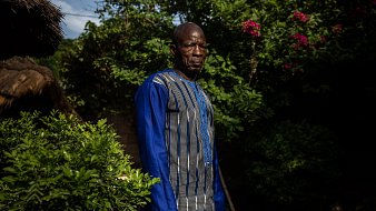 L'image montre un homme debout dans un environnement verdoyant. Il porte une tunique à manches longues, qui est ornée de lignes bleues et de motifs subtils. Son expression est sérieuse et son regard est dirigé vers l'objectif. Autour de lui, on peut apercevoir des plantes luxuriantes, avec des feuilles brillantes et quelques fleurs colorées en arrière-plan. La lumière du soleil filtre à travers les branches, créant une atmosphère tranquille.
