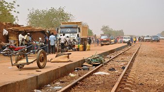 L'image dépeint une scène animée le long d'une voie ferrée. Sur la droite, des rails en métal s'étendent, entourés de poussière. À proximité, on aperçoit plusieurs véhicules, notamment des camions et des voitures, stationnés ou en mouvement. Des personnes se regroupent sur le côté, certains discutant, d'autres transportant des marchandises. Sur le sol, on peut distinguer divers objets laissés là, comme des déchets, des morceaux de métal et des câbles. À gauche, une petite charrette à deux roues est partiellement visible. En arrière-plan, la végétation est clairsemée, avec quelques arbres. Le ciel est d'un ton brumeux, suggérant une chaleur intense et un environnement rural. L'atmosphère générale est à la fois occupée et rustique, évoquant l'activité quotidienne dans une région où la vie s'organise autour des transports.