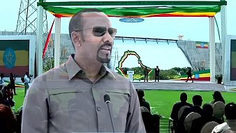 L'image montre un homme debout au premier plan, portant des lunettes de soleil et un uniforme militaire, qui semble s'adresser à un public. Il est probablement en train de faire un discours. En arrière-plan, on aperçoit une grande scène décorée de drapeaux éthiopiens et d'autres couleurs vives, avec une grande structure ressemblant à un barrage. Au premier plan, on peut voir des personnes assises, attentives, apprêtées pour un événement officiel en plein air, sur une pelouse verte. L'atmosphère semble d'une grande importance, peut-être liée à une célébration ou une inauguration.