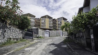 L'image montre une rue en pente, bordée de murs en pierre et de verdure. Sur le côté, on peut distinguer des fleurs colorées et des plantes tropicales. À l'arrière-plan, plusieurs bâtiments modernes avec des balcons se dressent, peints dans des teintes de jaune et de beige. Le ciel est nuageux, ce qui donne une ambiance légèrement dramatique à la scène. Le sol de la rue est inégal et montre des signes d'usure, avec des pavés qui apportent une texture variée au passage. L'atmosphère est calme, typique d'un quartier résidentiel.