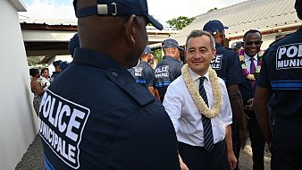 Dans cette image, nous voyons un groupe de policiers municipaux, tous vêtus d'un uniforme bleu avec le mot "POLICE" inscrit sur le dos. Au premier plan, un homme souriant échange une poignée de main avec un policier. Cet homme porte une chemise blanche et une cravate, ainsi qu'un collier de fleurs autour du cou, ce qui suggère un événement formel ou culturel. En arrière-plan, on aperçoit d'autres personnes, dont certaines semblent également habillées de manière soignée. La scène se déroule dans un environnement extérieur, avec des bâtiments aux toits en tôle. L'atmosphère semble accueillante et amicale.