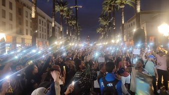 L'image montre une grande foule rassemblée dans une rue animée, éclairée par des lampadaires et entourée de palmiers. La scène se déroule en soirée, avec un ciel crépusculaire. Beaucoup de personnes tiennent des téléphones ou des lumières, créant un effet lumineux impressionnant. Les gens semblent mobilisés, certains tenant des pancartes et s'exprimant avec passion. L'atmosphère est énergique et engagée, reflétant une ambiance de manifestation ou d'événement public où les participants cherchent à faire entendre leur voix.