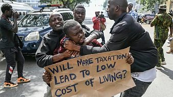 L'image montre une scène de protestation urbaine. Au premier plan, deux hommes sont engagés dans une altercation. L'un d'eux semble essayer de saisir l'autre qui tient une pancarte. Sur cette pancarte, on peut lire une question écrite en lettres noires : "WILL MORE TAXATION LEAD TO LOW COST OF LIVING?" (Plus de taxation conduira-t-elle à un coût de la vie plus bas ?). En arrière-plan, on aperçoit d'autres personnes, certaines les regardent, tandis que d'autres semblent prendre des photos. Des véhicules sont garés sur le côté, ajoutant à l'ambiance animée de la scène. Les expressions des hommes montrent des émotions vives, reflétant l'intensité de la situation.