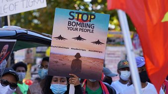 L'image montre une scène de manifestation. Au premier plan, une personne tient une grande pancarte qui affiche le message "STOP BOMBING YOUR PEOPLE" avec des images d'avions de chasse en vol. La personne est entourée d'autres manifestants portant des masques faciaux, certains brandissant d'autres pancartes. En arrière-plan, on peut voir des drapeaux et des affiches qui expriment des messages de protestation. L'atmosphère est chargée d'émotion, soulignant un appel urgent à l'action.