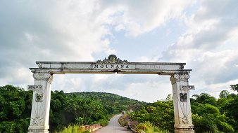 L'image présente une grande arche située à l'entrée de la ville de Mounana. Cette arche, ornée de motifs architecturaux, est peinte en blanc avec des détails dorés et armoriaux de chaque côté. Au-dessus de l'entrée, le nom "MOUNANA" est inscrit en lettres stylisées. En arrière-plan, on aperçoit un paysage verdoyant avec des collines et une route qui serpente entre des arbres. Le ciel est partiellement nuageux, créant une atmosphère paisible et accueillante. L'ensemble évoque l'entrée d'un lieu significatif, invitant les visiteurs à découvrir ce qui se trouve au-delà de l'arche.