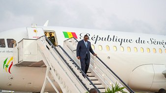L'image montre un homme en costume sombre descendant d'un avion. L'avion, aux couleurs blanches et avec l'inscription "République du Sénégal" sur le fuselage, est garé sur une piste. À l'arrière-plan, le ciel est légèrement nuageux. Le personnage semble marcher avec assurance, et des escaliers mènent directement à la porte de l'avion. Au sol, on peut voir quelques plantes, suggérant un environnement accueillant.