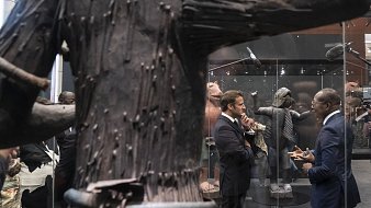 L'image montre un espace d'exposition où deux hommes en costume discutent. L'un d'eux semble concentré sur la conversation, tandis que l'autre écoute attentivement. Ils se tiennent près d'une grande sculpture en bois, qui occupe une place centrale. Cette sculpture a une texture rugueuse, avec des détails qui évoquent une forme humaine ou abstraite. En arrière-plan, d'autres œuvres d'art sont visibles, ajoutant à l'atmosphère culturelle de l'exposition. L'éclairage est doux, mettant en valeur les sculptures tout en créant une ambiance contemplative et respectueuse.