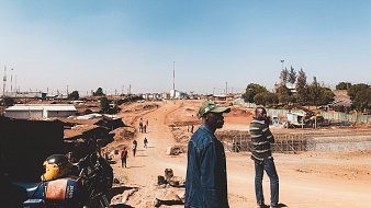 Dans cette image, nous voyons une scène d'une rue en construction dans une zone urbaine. Le ciel est dégagé et d'un bleu clair. Au premier plan, deux hommes se tiennent debout : l'un porte une veste en jean et une casquette, tandis que l'autre est vêtu d'un sweater rayé. À droite, on aperçoit un chemin de terre, entouré de bâtiments en dur et d'habitations simples. À gauche, une moto est garée, et plusieurs personnes se déplacent le long de la route. L'atmosphère semble calme, mais l'environnement indique une activité en cours avec des travaux de construction en arrière-plan.
