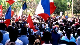 L'image représente une foule dense lors d'un événement, probablement un rassemblement ou une manifestation. Au premier plan, on peut voir des personnes brandissant des drapeaux français, avec des couleurs bleu, blanc et rouge. Certains membres de la foule portent des maquillages colorés sur le visage, ajoutant une dimension festive ou militante à la scène. Les visages sont à la fois heureux et engagés, témoignant d'une ambiance dynamique. En arrière-plan, des arbres et de la verdure suggèrent que l'événement se déroule dans un espace extérieur, peut-être dans un endroit tropical ou rural. L'ensemble dégage une forte impression de solidarité et de passion collective.