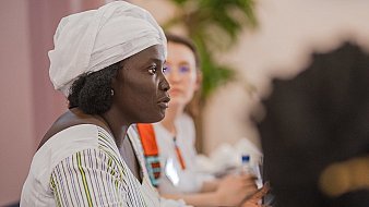 L'image montre une femme assise à une table lors d'une réunion ou d'une discussion. Elle porte une tenue traditionnelle, avec une chemise à rayures et un foulard blanc sur la tête. Son expression est sérieuse et concentrée, et elle semble engagée dans la conversation. À côté d'elle, une autre femme est présente, mais elle est floue en arrière-plan. On peut apercevoir des bouteilles d'eau sur la table, ainsi qu'une ambiance chaleureuse et collaborative. Des éléments de verdure, comme une plante, ajoutent une touche de couleur à la scène.