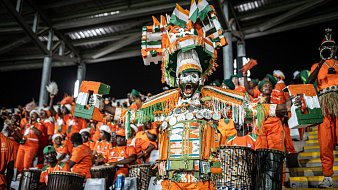 L'image montre une scène vibrante d'un événement festif, probablement un match de football. Au premier plan, un homme se distingue par son costume coloré composé de tissus orange et de nombreux accessoires décoratifs, dont des coquillages et des éléments en métal qui brillent à la lumière. Il est entouré de groupes de personnes, également en orange, qui jouent des tambours et sont vêtus de vêtements similaires. Tous semblent animés et enthousiastes, contribuant à une atmosphère de fête collective, ponctuée de chants et de danses. Les visages sont exprimés par des sourires et de la joie, avec des éléments traditionnels mettant en avant la culture locale.