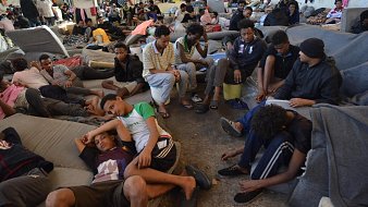 L'image montre un grand espace intérieur, probablement une salle ou un centre d'accueil, où de nombreuses personnes sont rassemblées. Le sol est recouvert de matelas ou de couvertures, et les gens sont assis ou allongés, souvent en petits groupes ou seuls. Les visages montrent une variété d'émotions, allant de la fatigue à l'attente. L'atmosphère semble chargée, et l'agencement des personnes donne une idée d'une situation difficile. Les murs sont nus, et il y a des effets personnels éparpillés ici et là. L'environnement est sombre et témoigne d'un manque de confort.