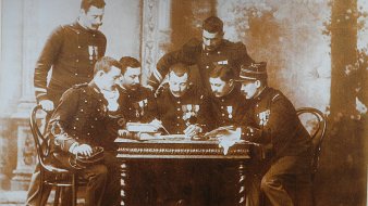 Cette image en noir et blanc représente un groupe de six hommes en uniforme, assis autour d'une table en bois. Ils semblent être des membres d'un corps militaire, chacun portant des médailles décoratives sur leur uniforme. Les hommes sont concentrés sur un document ou un plan posé sur la table, et leur posture indique un échange d'idées ou de discussions sérieuses. En arrière-plan, on peut distinguer quelques éléments architecturaux flous, suggérant un intérieur de bâtiment classique. L'atmosphère se dégage une certaine solennité et camaraderie.