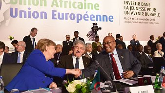 L'image montre la scène d'une conférence internationale où des dirigeants participent à un sommet. Au premier plan, on aperçoit un groupe de personnes, parmi lesquelles une femme, identifiée comme une figure politique importante, est en train de serrer la main d'un homme. Cette interaction symbolise le dialogue et la collaboration entre les nations. À l'arrière-plan, on distingue des bannières et des affiches portant le titre "Union Africaine, Union Européenne" ainsi que le slogan "Investir dans la jeunesse pour un avenir durable". La salle semble remplie de délégués, en costard et concentrés sur les discussions, ce qui donne une atmosphère formelle et engagée. Des éléments comme une table avec des drapeaux de différents pays ajoutent au cadre diplomatique de l'événement.