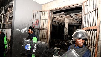 L'image montre une scène de sécurité, probablement après un événement perturbateur. On y voit deux agents de sécurité, vêtus d'un uniforme bleu et vert, portant des casques de protection et tenant des boucliers. Ils se tiennent devant une entrée, à côté de portes métalliques ouvertes. À l'intérieur, on aperçoit un espace sombre avec des traces de dégâts, comme des murs noircis. L'environnement semble chaotique, avec des débris visibles au sol. Des graffitis sont présents sur les murs, ajoutant une touche de désordre à la scène. L'atmosphère générale dégage un sentiment de tension et de vigilance.